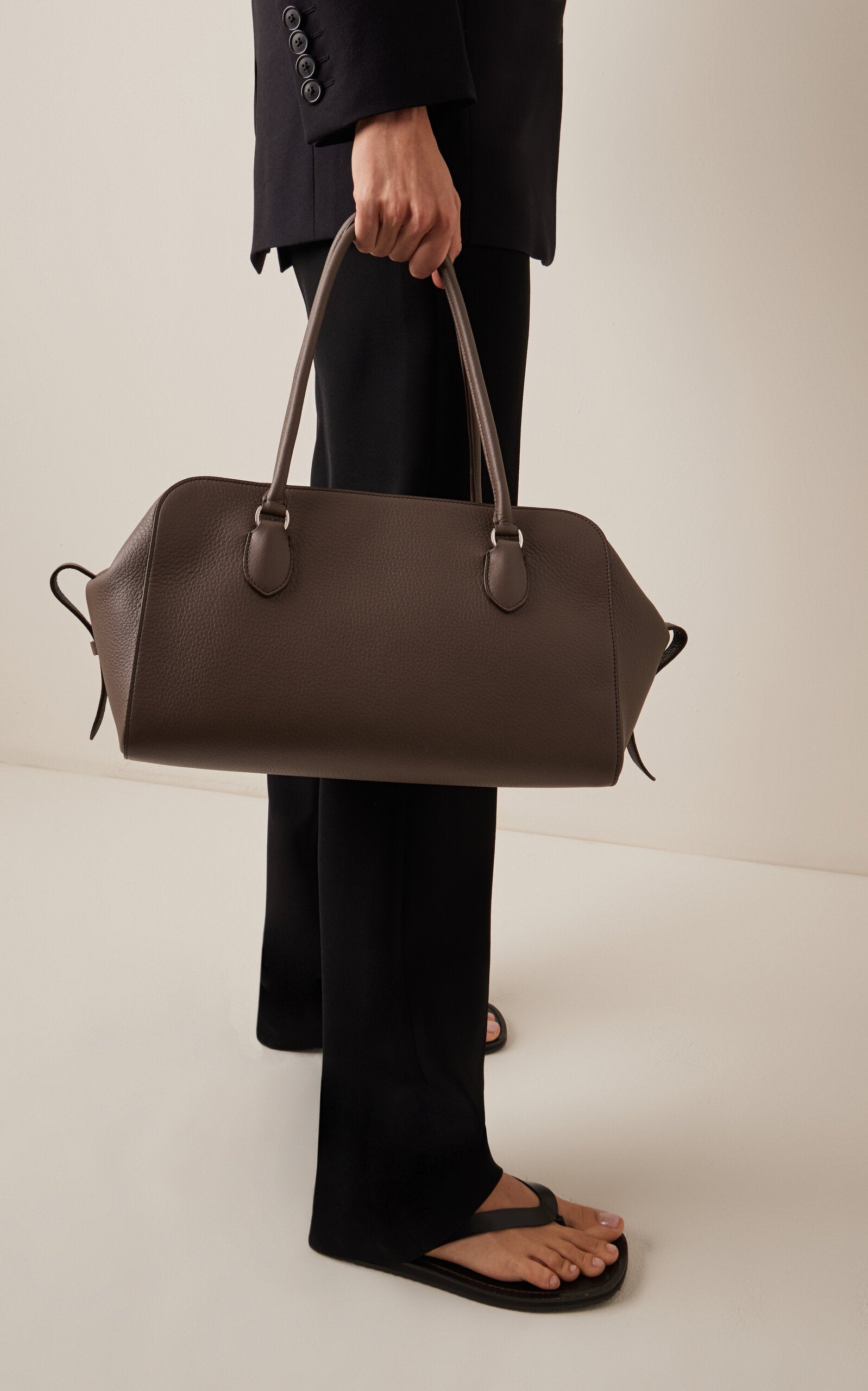 Crafted from luxurious grained calfskin leather, the E/W Leather Bowling Bag by The Row combines refined simplicity with architectural precision. Its structured silhouette features distinctive gussets and sturdy tubular handles, providing both style and durability. The secure zip-top closure ensures your essentials remain safe, making this bag a timeless accessory that embodies meticulous craftsmanship and understated elegance.