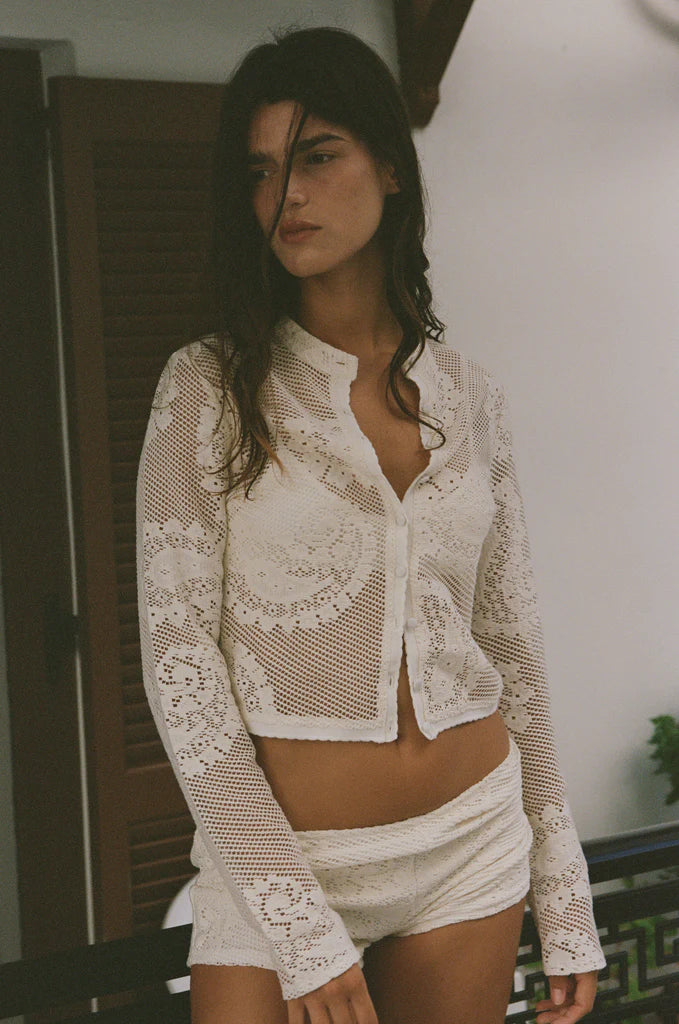 The Dom Top by With Jean is an expertly crafted long-sleeve cardigan with a button-up front and a refined neckline collar. It features delicate scalloped trim and custom hand-drawn crochet lace detailing, combining intricate craftsmanship with timeless style for a sophisticated and elegant look.