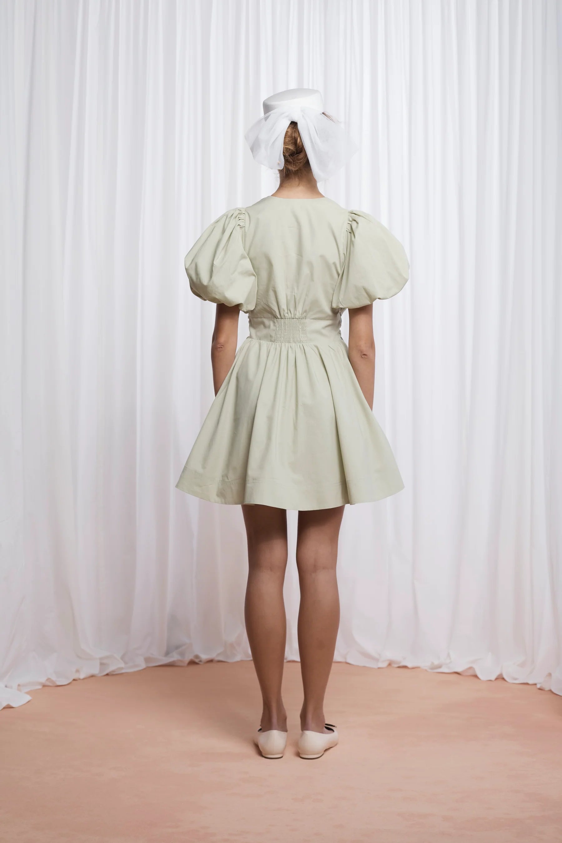 The Devon Mini Dress in pistachio combines expert tailoring with comfort, crafted from butter-soft cotton poplin. Its deep V-neckline and blousy puff sleeves create a flattering silhouette, while the double bow front adds a refined, feminine touch. Designed for ease, it features an elasticated back, discreet side seam zip, and practical pockets. Fully lined for smooth wear, this dress delivers both style and function in a lightweight, breathable fabric.