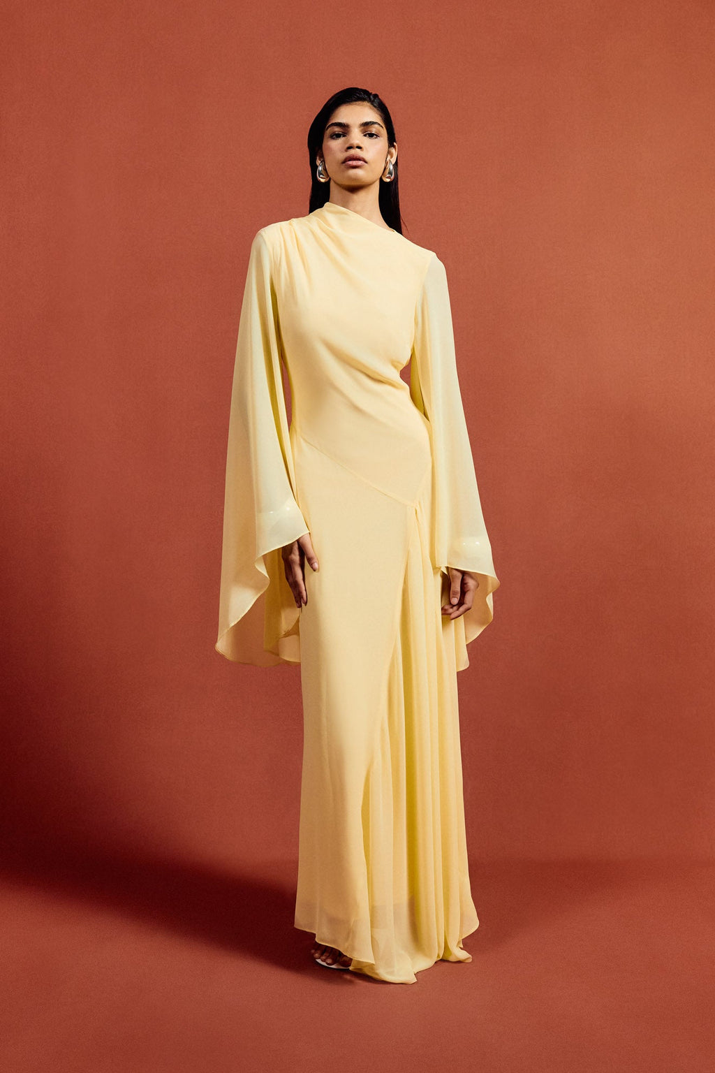 The Hausos Maxi Dress in Sherbet Yellow features an elegant chiffon construction with a floor-length silhouette, creating a graceful presence. Its distinctive dipped waist seam and draped skirt with godet panels add dynamic movement and refined shape. The asymmetric neckline delivers a modern edge, while the long pagoda sleeves introduce dramatic flare for a sophisticated, fashion-forward statement. Ideal for formal occasions, this dress combines fluidity and structure with expert design details.