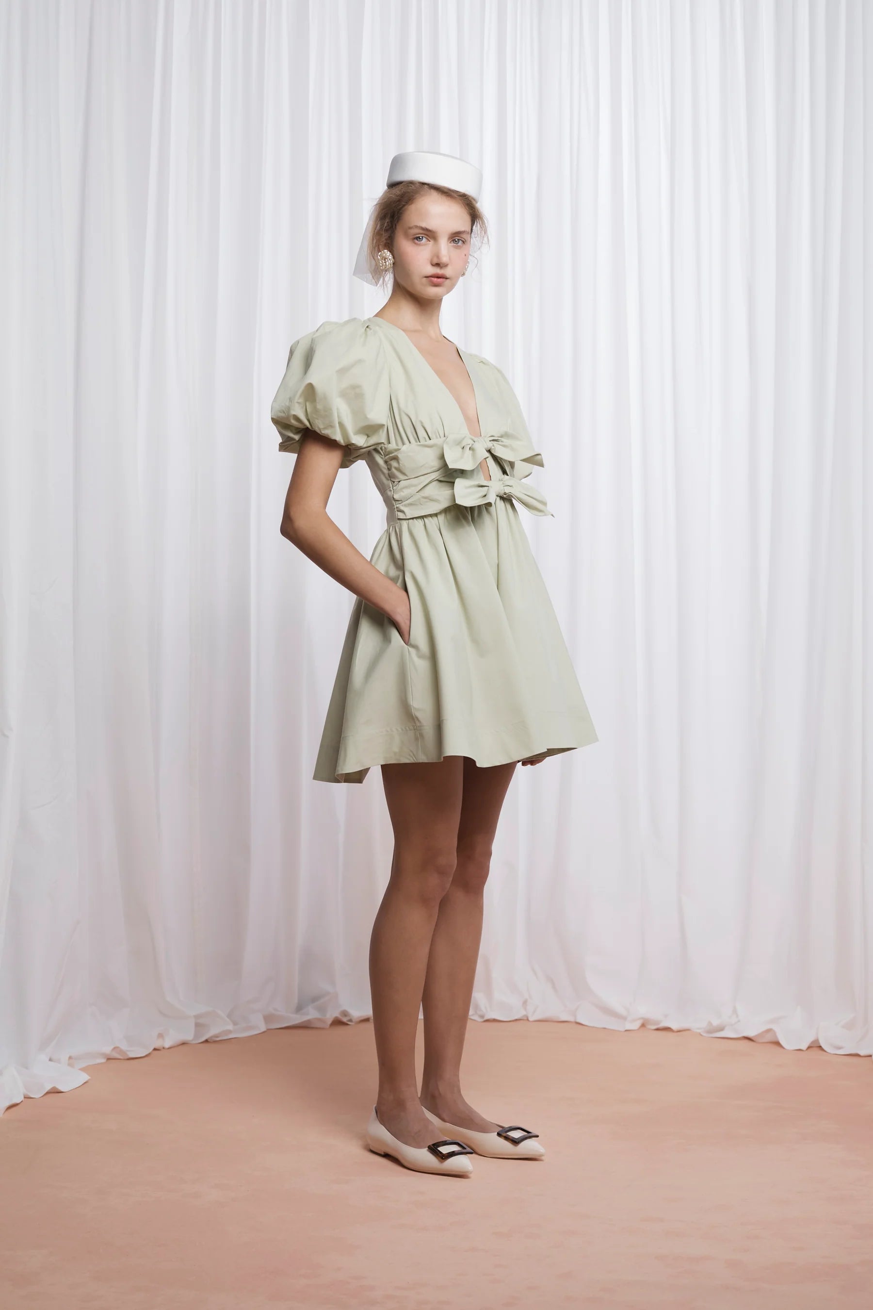 The Devon Mini Dress in pistachio combines expert tailoring with comfort, crafted from butter-soft cotton poplin. Its deep V-neckline and blousy puff sleeves create a flattering silhouette, while the double bow front adds a refined, feminine touch. Designed for ease, it features an elasticated back, discreet side seam zip, and practical pockets. Fully lined for smooth wear, this dress delivers both style and function in a lightweight, breathable fabric.