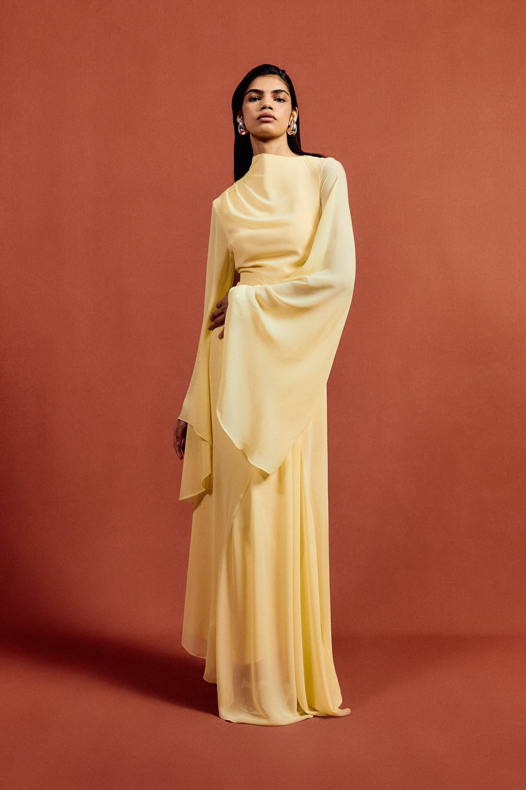 The Hausos Maxi Dress in Sherbet Yellow features an elegant chiffon construction with a floor-length silhouette, creating a graceful presence. Its distinctive dipped waist seam and draped skirt with godet panels add dynamic movement and refined shape. The asymmetric neckline delivers a modern edge, while the long pagoda sleeves introduce dramatic flare for a sophisticated, fashion-forward statement. Ideal for formal occasions, this dress combines fluidity and structure with expert design details.